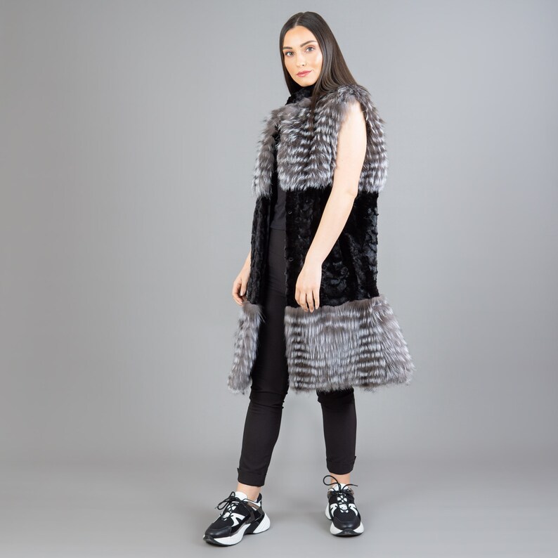 A female model is posing in front of a gray panel. She is wearing a vest made from real fox and mink fur in black and silver colors.