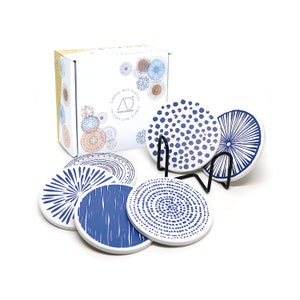 May include: Set of six blue and white ceramic coasters with different geometric patterns. The coasters are displayed on a black metal stand. The box for the coasters is in the background and says "Simple, But Special".