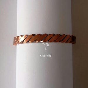 Può includere: Un braccialetto color rame con un design a corda intrecciata. Il braccialetto è in metallo e ha una finitura lucida. Il braccialetto è esposto su uno sfondo bianco. Il testo "Kikansie" è nell'immagine.