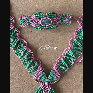 May include: A pink and green macrame necklace and bracelet set with a green gemstone pendant. The necklace features a teardrop-shaped pendant with a green gemstone in the center. The bracelet is a simple band with a green gemstone in the center. The set is made with macrame knots and beads.