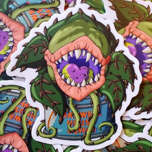 May include: A sticker featuring a cartoon illustration of a green, toothy plant with a large mouth, eating a purple and green object. The plant is growing out of a blue and orange can labeled "Maxwell House Coffee".