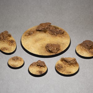 Puede incluir: Conjunto de bases de terreno en miniatura con temática desértica. Las bases tienen varias formas y tamaños, con una combinación de colores beige y marrón. Cada base presenta detalles texturizados y pequeñas formaciones rocosas.