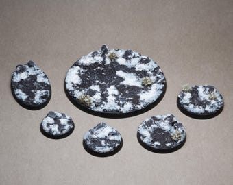 May include: A collection of miniature terrain bases, featuring a textured black and white design resembling snow-covered ground. The bases vary in size and shape, with small tufts of dried vegetation.