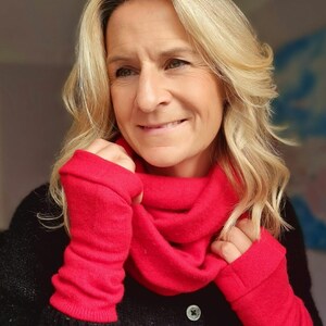 Könnte beinhalten: Eine Frau trägt einen roten Strickschal und passende fingerlose Handschuhe. Die Frau trägt einen schwarzen Pullover mit Knopfverschluss. Der Hintergrund ist verschwommen, mit Andeutungen von Blau und Weiß.