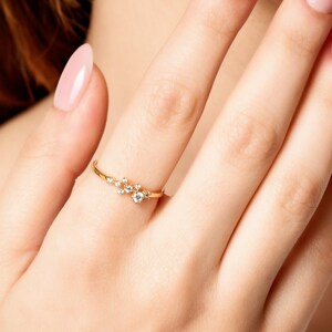 Pu&ograve; includere: Un delicato anello d'oro con un gruppo di piccole pietre chiare incastonate in un design delicato e curvo.