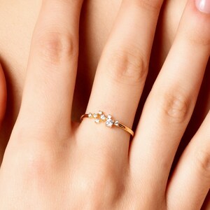 Pu&ograve; includere: Un anello d'oro con un gruppo di piccoli diamanti incastonati in una fascia delicata. L'anello &egrave; indossato sull'anulare di una mano con smalto per unghie rosa chiaro.