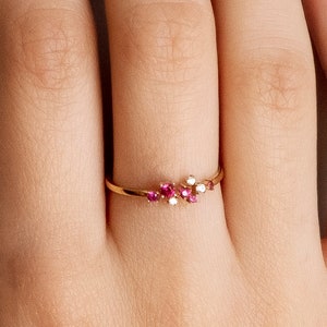 Könnte beinhalten: Ein zarter Goldring mit einem Cluster aus kleinen rosafarbenen Edelsteinen, die in einer geschwungenen Linie angeordnet sind.