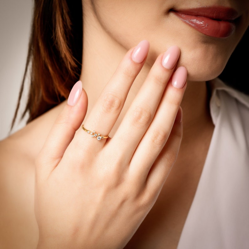 Pu&ograve; includere: Un delicato anello d'oro con un gruppo di piccole pietre chiare incastonate in un design delicato e curvo.