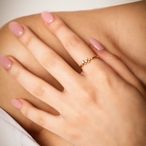 Pu&ograve; includere: Un delicato anello d'oro con un gruppo di piccole pietre preziose trasparenti. L'anello &egrave; indossato sul dito di una mano con smalto per unghie rosa chiaro.