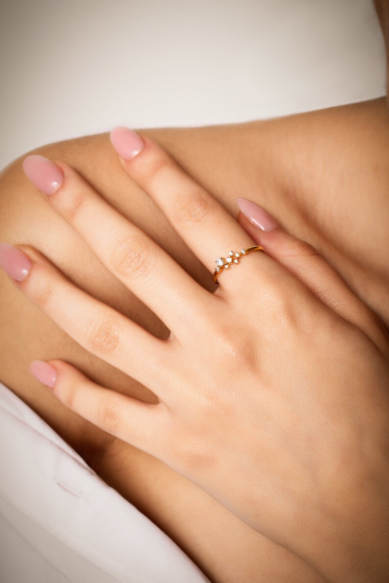 Pu&ograve; includere: Un delicato anello d'oro con un gruppo di piccole pietre preziose trasparenti. L'anello &egrave; indossato sul dito di una mano con smalto per unghie rosa chiaro.
