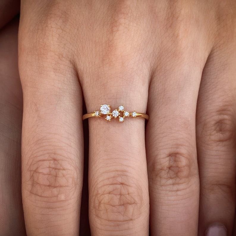 Pu&ograve; includere: Un delicato anello d'oro con un gruppo di piccoli diamanti incastonati in un design minimalista.