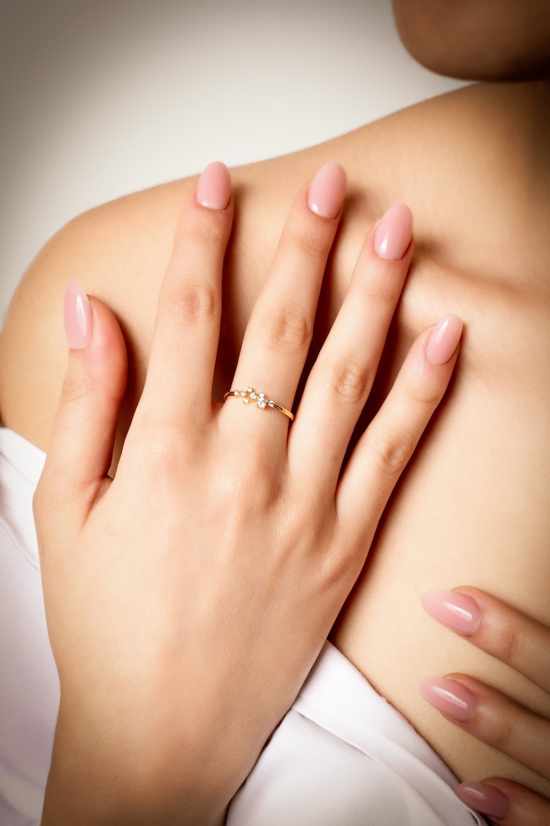 Pu&ograve; includere: Un anello d'oro con un gruppo di piccoli diamanti incastonati in una fascia delicata. L'anello &egrave; indossato sull'anulare di una mano con smalto per unghie rosa chiaro.