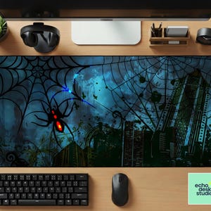 May include: A large rectangular desk mat featuring a dark blue and teal spiderweb design with a large black spider with red eyes. The background includes city buildings and a teal and blue sky. The mat has the text "echo desk studio".