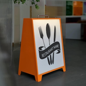 以下が含まれることがあります： フォーク、スプーン、ナイフのグラフィックが描かれた白いパネルが付いた、オレンジ色のA型看板。黒いバナーには「RESTAURANT NAME」と表示されています。この看板には銀色のハンドルが付いており、レストランに最適です。