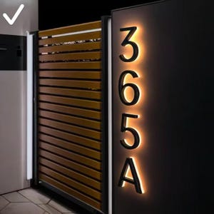 May include: Modern illuminated house numbers 365A in a sleek black font, mounted vertically on a dark panel. The numbers are backlit with a warm, inviting glow. A wooden gate is visible to the left.