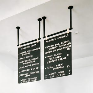 Könnte beinhalten: Zwei schwarze rechteckige Menütafeln, die von der Decke hängen, mit schwarzen Stangen und weißem Text. Die linke Tafel zeigt "Baguette Menu" mit Menüpunkten. Die rechte Tafel zeigt "Maison's Specials" mit Menüpunkten und Preisen.