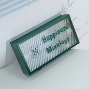 May include: Green rectangular sign with the words "Happiness Mixology" in white lettering. The sign has a silver, textured background and a small circle with Chinese characters in the centre.