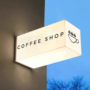 Puede incluir: Un letrero rectangular blanco con las palabras "COFFEE SHOP" en texto negro. El letrero tiene una silueta negra de una taza de café con vapor saliendo de ella.