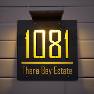 May include: A black rectangular address sign displaying the number "1081" in a bright yellow font. The text "Thara Bey Estate" is below the numbers, also in yellow. The sign is mounted on a wall.