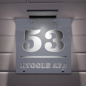 May include: A contemporary silver address sign displaying the number "53" in a cut-out design, backlit for illumination. The street name "KYOGLE AVE" is also cut out below. The sign is mounted on a light-toned wall.