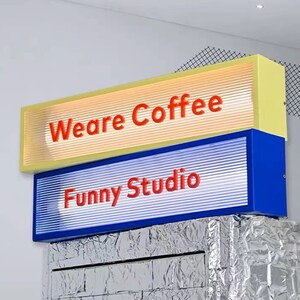 May include: Two illuminated signs with a yellow and blue frame. The top sign reads "We are Coffee" in red lettering. The bottom sign reads "Funny Studio" in red lettering.
