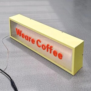 May include: A yellow rectangular sign with the words "We are Coffee" in red lettering. The sign is illuminated from behind, creating a soft glow.