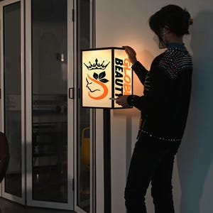 May include: A lit sign with the words "GLOBAL BEAUTY" in orange and a logo featuring a crown, leaves, and a woman's profile. The sign is mounted on a black pole.