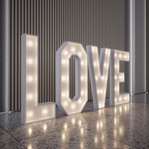 May include: Large white marquee letters spelling "LOVE" with illuminated bulbs. The letters are standing on a speckled floor, with a textured wall in the background. The lights are on, creating a warm glow.
