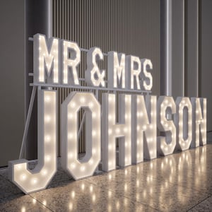 May include: White marquee letters spelling "MR & MRS JOHNSON" with warm white lights. The letters are freestanding and arranged on a reflective surface, ideal for wedding or event decor. The letters are illuminated, creating a bright and festive display.