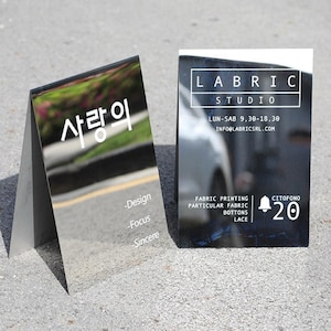 Könnte beinhalten: Zwei reflektierende Visitenkarten. Eine Karte hat koreanischen Text und die Wörter "Design, Focus, Sincere". Die andere Karte hat den Text "LABRIC STUDIO" und Geschäftszeiten. Die Karten liegen auf einer grauen Oberfläche.
