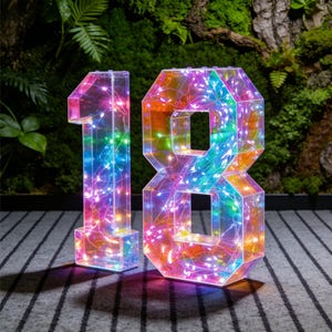 Puede incluir: Un número 18 geométrico transparente iluminado con luces LED de colores. Los números están hechos de material transparente, lo que permite que las luces brillen, creando un vibrante efecto arcoíris. Ideal para celebraciones de cumpleaños o eventos importantes.