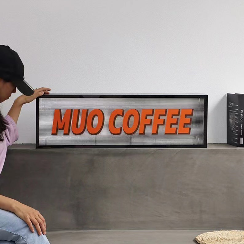 May include: A black framed sign with the words "MUO COFFEE" in orange letters. The sign is sitting on a gray surface.