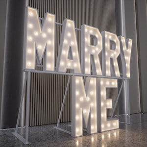 May include: A large, illuminated sign spells out "MARRY ME" in white block letters. Each letter is outlined with warm, glowing light bulbs. The sign is supported by a white metal frame, perfect for a wedding or proposal.