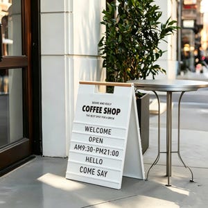 May include: A-frame sign for a coffee shop with the text "COFFEE SHOP" and "THE BEST SPOT FOR A BREW". The sign also says "WELCOME", "OPEN", "AM9:30-PM21:00", "HELLO", and "COME SAY". A small table and plant are in the background.