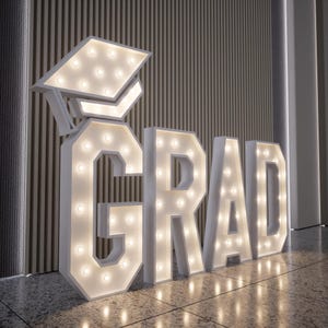 May include: Large white marquee letters spelling "GRAD" with a graduation cap, illuminated by warm white lights. The letters are placed on a tiled floor, perfect for graduation celebrations and photo opportunities.