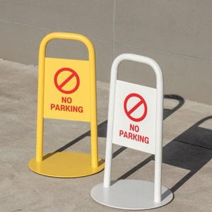 Puede incluir: Dos señales metálicas de "Prohibido estacionar". Una es amarilla con un círculo rojo y un símbolo de barra, y las palabras "NO PARKING" en rojo. La otra es blanca con el mismo símbolo y texto. Ambas tienen una parte superior redondeada y una base circular.