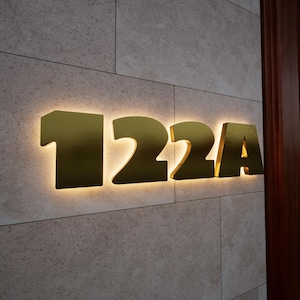 May include: Gold-colored house numbers, "122A", are mounted on a textured, light-colored wall. The numbers are illuminated from behind, creating a warm glow. The wall is made of rectangular tiles.