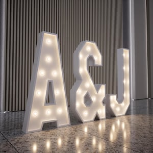 Op de afbeelding: Witte marquee letters A, &, en J, verlicht met warmwitte lampen. De letters staan op een gevlekte vloer, geplaatst voor een verticale houten wand. De verlichting zorgt voor een zachte gloed.