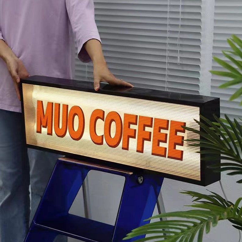 May include: A black rectangular sign with a white background and orange text that reads "MUO COFFEE". The sign is illuminated from behind.