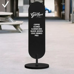 May include: A black, oval-shaped sandwich board sign with the text "The Good Spot Coffee & More" in white script. Below, the sign lists "Coffee, Scones, Sandwiches, Baked Goods, Cold Drinks, Pies." The sign is mounted on a black metal stand.