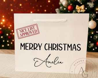 May include: White gift bag with the text "MERRY CHRISTMAS Amelia" in black, and a red stamp that says "NICE LIST APPROVED". The bag has a white rope handle and a logo in the bottom right corner.