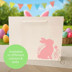 May include: A cream gift bag featuring a pink bunny silhouette and the name "Isabella". The bag has a white handle and the text "Available in different colours & sizes" in a green circle. Easter-themed background.