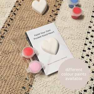 May include: A craft kit with the text "Paint Your Own Pocket-Sized Heart" includes a heart-shaped item, paint pots in red, pink, and blue, and a paintbrush. The kit is designed for creative activities and comes with different color paints.