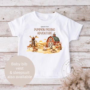 May include: White toddler t-shirt featuring a farm scene with a barn, windmill, and animals, and the text "Spencer's First Pumpkin Picking Adventure." Baby bib, vest, and sleepsuit also available. The design is in autumnal colours.