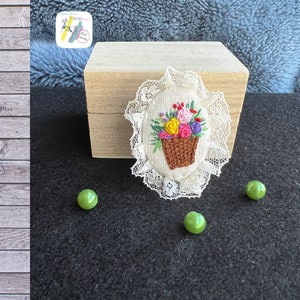 Op de afbeelding: Een witte ovale broche met een kantversiering, met een geborduurde mand met kleurrijke bloemen. De broche ligt op een donker oppervlak met drie groene kralen.