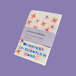 May include: A small white notebook with a pink floral pattern and the text "coming-out heftchen" in black. The text below reads "für non-binarys, trans*, agender und und und". The notebook also has the text "WUSSTEST DU EIGENTLICH DASS..." in blue.
