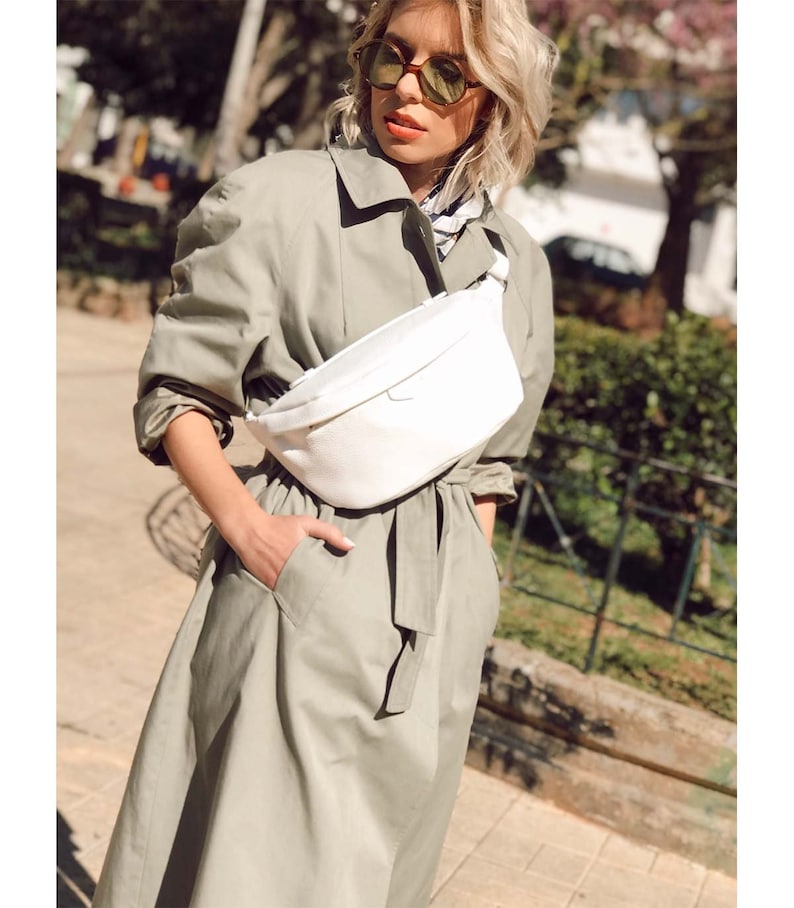 May include: A woman wearing a light green trench coat with a white fanny pack worn around her waist. She is wearing sunglasses and has blonde hair.