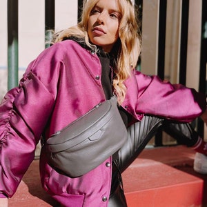 May include: A woman wearing a pink satin bomber jacket with black buttons and black leather pants. She is wearing a gray leather fanny pack with a zipper.
