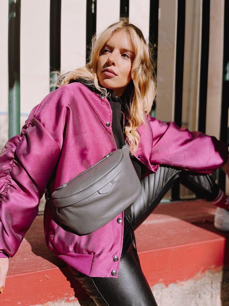 May include: A woman wearing a pink satin bomber jacket with black buttons and black leather pants. She is wearing a gray leather fanny pack with a zipper.