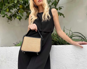May include: A black sleeveless dress with a side slit. The person is holding a small, rectangular handbag with a woven straw front and black trim. The person is wearing orange-tinted sunglasses.